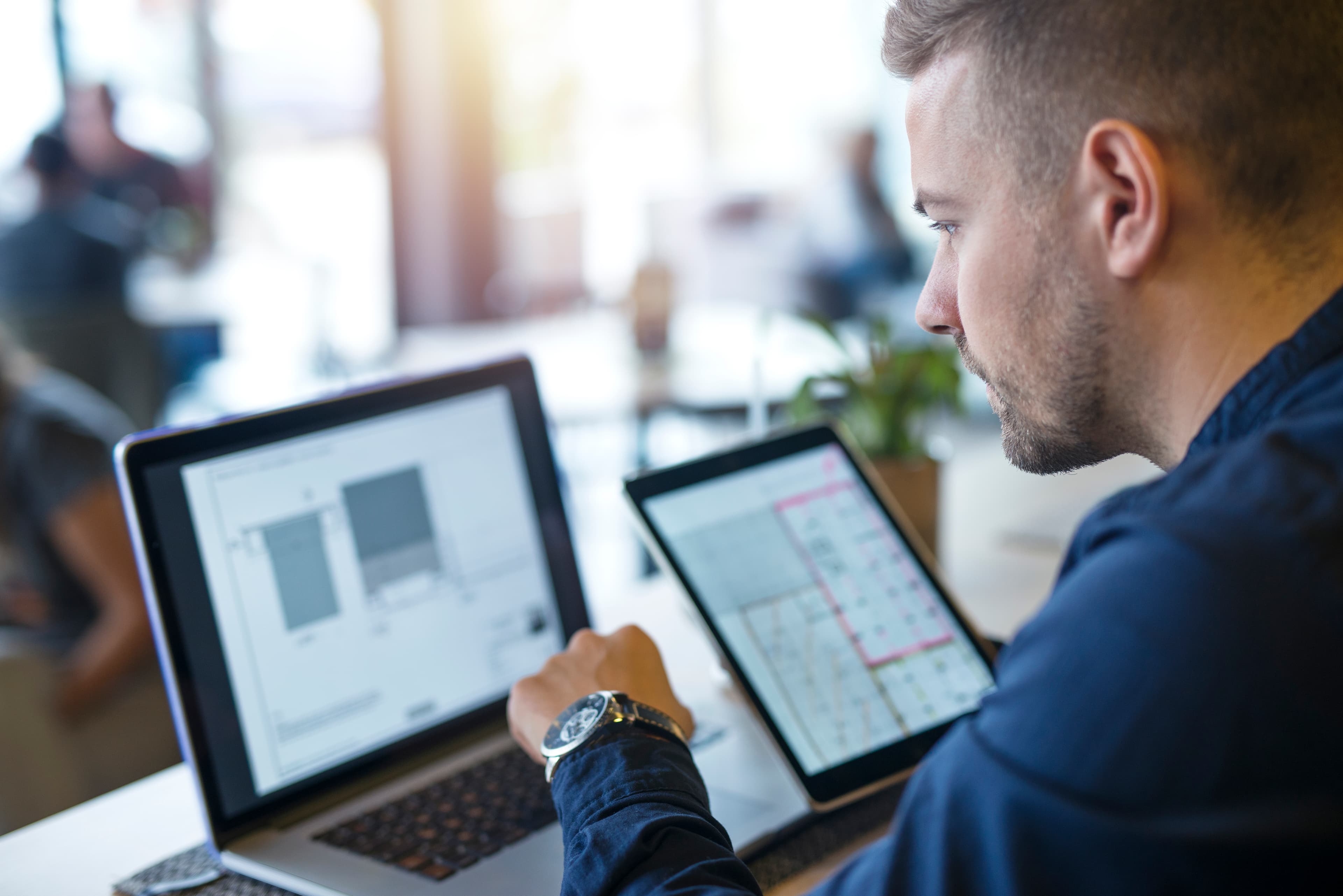 Homem de perfil concentra-se em um notebook e um tablet com diagramas, em um ambiente claro.